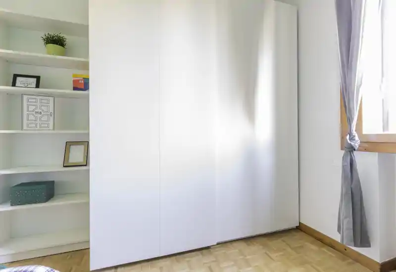 The photo shows a built-in wardrobe/closet with sliding white doors, open shelving to the left with decorative boxes and frames, parquet flooring and a curtained window to the right. No bed, sofa, kitchen fixtures or bathroom elements are visible — the image focuses on storage and a corner of the room.
