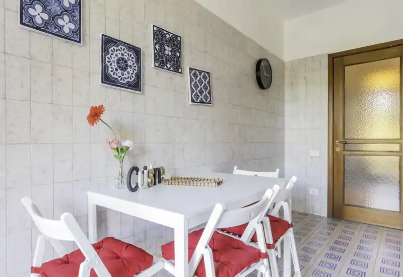Bright dining area with a white table and four folding chairs, red seat cushions, wall tiles and decorative framed tiles; simple, airy and well-styled for shared meals.