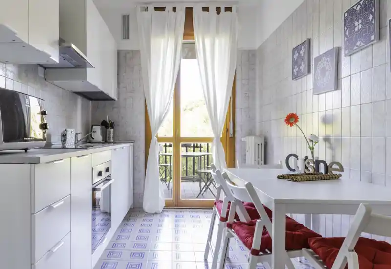 Bright modern kitchen with a small dining area: white cabinets, built-in oven, microwave and a table with red-cushioned chairs opening onto a sunny balcony — ideal for cozy breakfasts.