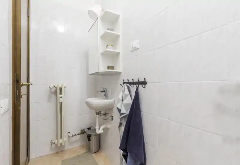 Compact white-tiled bathroom with a small wall-mounted sink, mirrored medicine cabinet and towel hooks. Clean, bright and efficiently arranged for a small washroom.