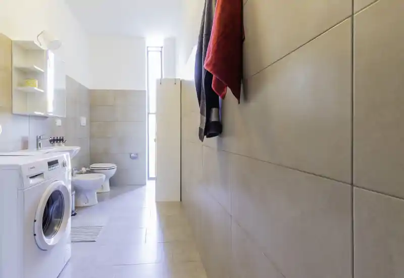 Clean, bright bathroom with a washing machine, sink, bidet and toilet; neutral tiles and natural light from a tall window create a fresh, functional space.
