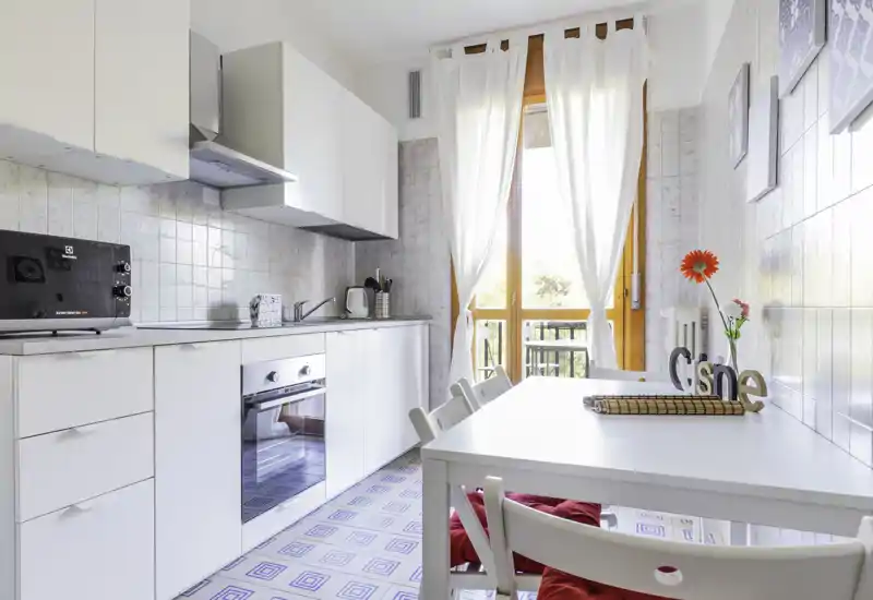 Bright modern kitchen with white cabinets, built-in oven, dining table and natural light from a balcony door — ideal for cozy breakfasts.