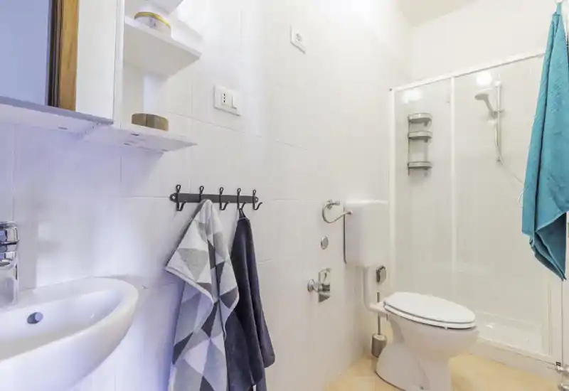Bright, clean bathroom with shower enclosure, toilet and wall-mounted sink; fresh towels on hooks and neutral tile walls for a welcoming, functional space.