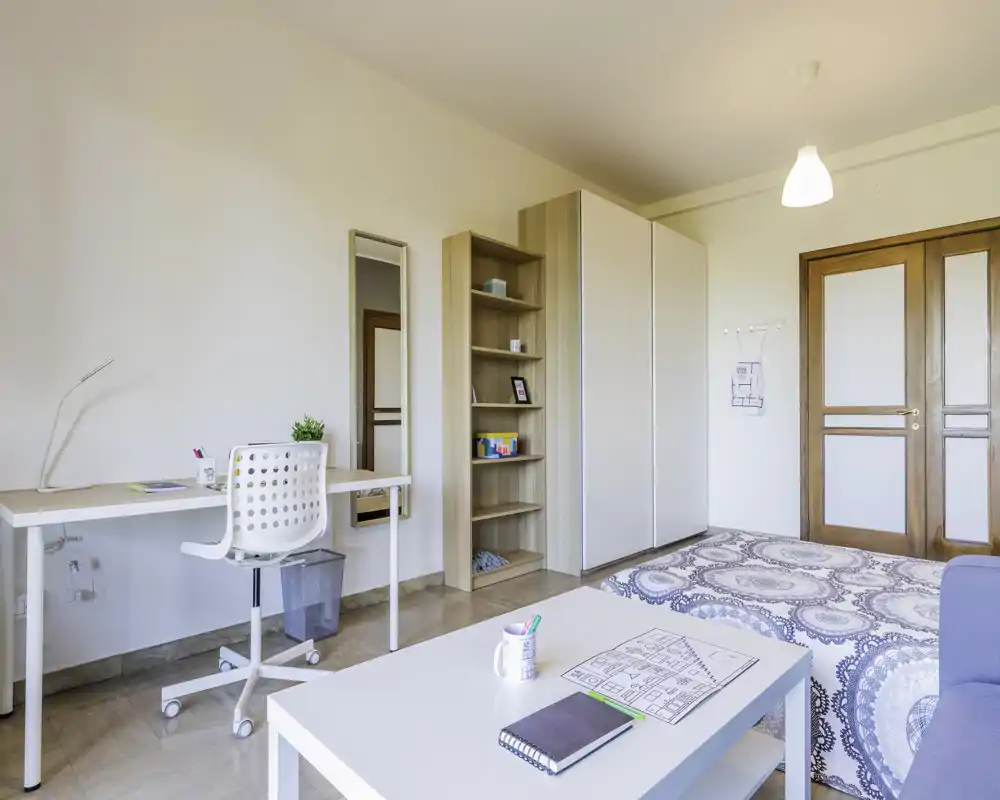 Camera da letto luminosa e ordinata con letto matrimoniale, scrivania e spazio contenitivo. Il soggiorno dispone di una coperta a motivo, libreria, armadio e una postazione di lavoro sulla parete — perfetta per riposo e smart working.
