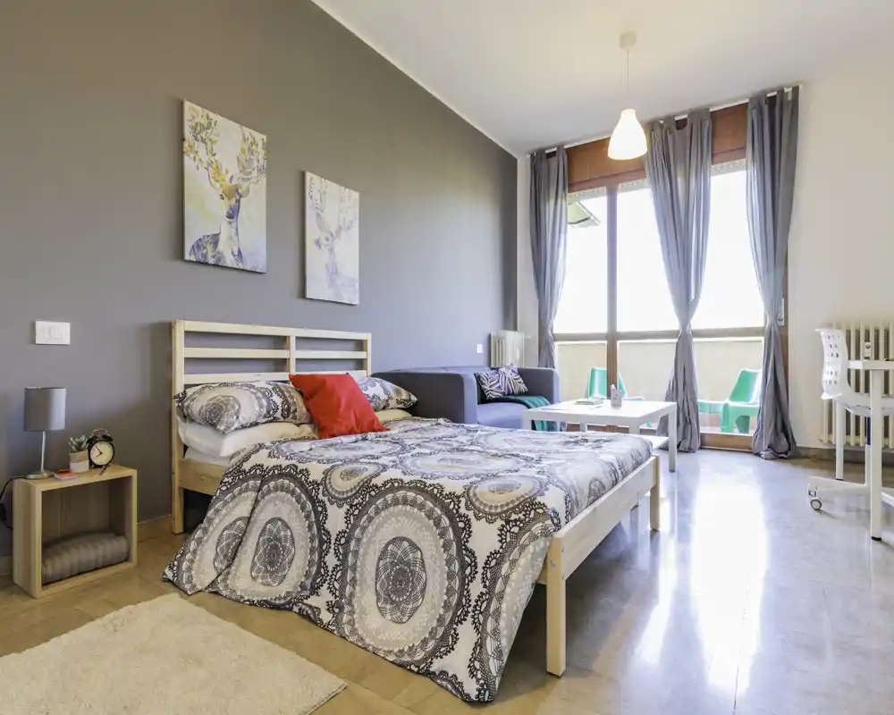 Camera da letto luminosa e moderna con un grande letto in primo piano, cuscini decorativi, un piccolo divano e una porta finestra che illumina l'ambiente con luce naturale.
