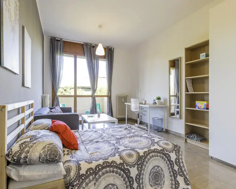 Camera da letto luminosa e ben arredata con letto matrimoniale in primo piano, piccolo salottino e scrivania vicino a una grande finestra che illumina l'ambiente.