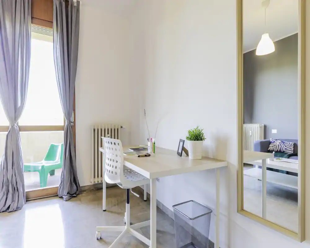 Zona giorno luminosa con scrivania e sedia vicino al balcone; stile moderno e minimale, pianta decorativa e divano visibile nello specchio creano un angolo studio nel soggiorno.