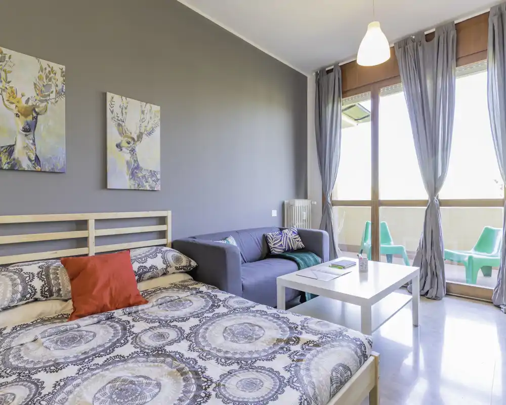 Camera da letto luminosa e ben arredata con letto matrimoniale, tessili accoglienti e un piccolo angolo salotto davanti alle ampie finestre e al balcone. Arredo moderno e pulito con luce naturale e quadri di buon gusto.