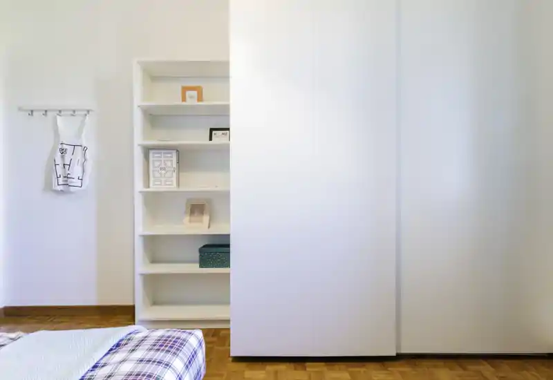 Angolo di camera minimalista con angolo di letto visibile, armadio a muro bianco e scaffalatura aperta con cornici decorative; luminoso, pulito e dallo stile semplice.