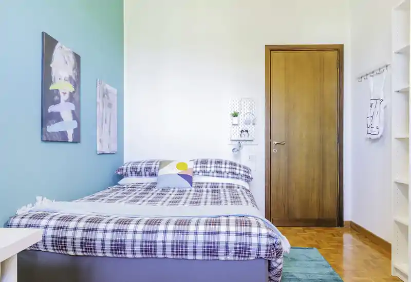 Camera da letto luminosa e ordinata con letto matrimoniale con lenzuola a quadri, cuscino d'accento colorato e parete turchese. Pavimento in parquet pulito e arredo minimalista che creano un'atmosfera moderna e accogliente.
