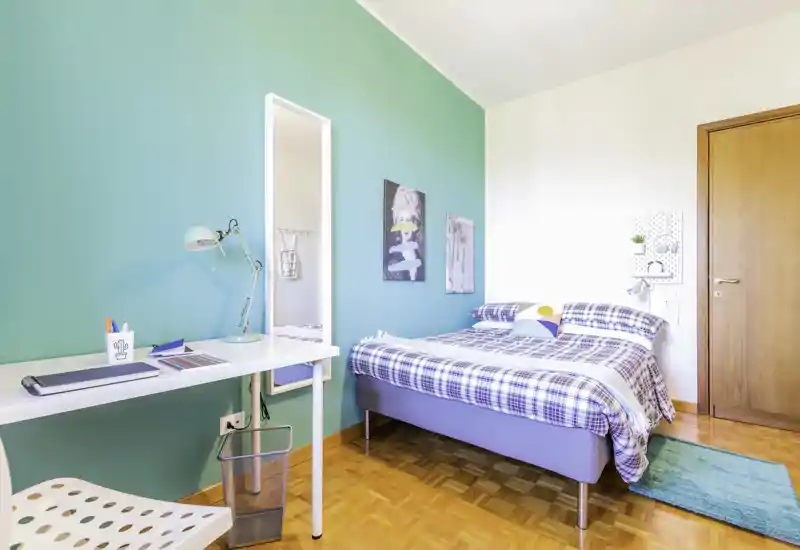 Camera da letto luminosa e moderna con letto matrimoniale in plaid, parete tinta turchese, scrivania e specchio a figura intera — ideale per studenti o soggiorni brevi.