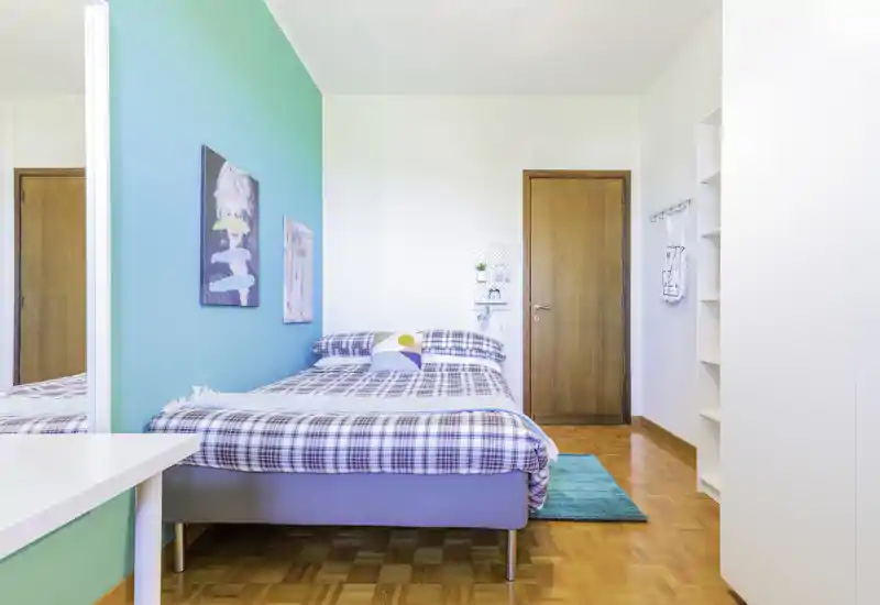 Camera da letto luminosa e ben allestita con letto matrimoniale contro una parete turchese, biancheria a quadri, cuscino decorativo e tappeto coordinato — perfetta per annunci che cercano una stanza moderna e vivace.