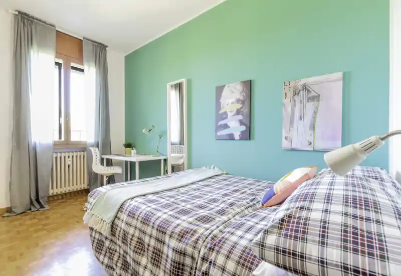 Camera da letto luminosa e arredata con letto ampio, copertura a quadri, parete accentuata color turchese, scrivania vicino alla finestra e quadri decorativi — perfetta per annunci che puntano su luce naturale e stile moderno.