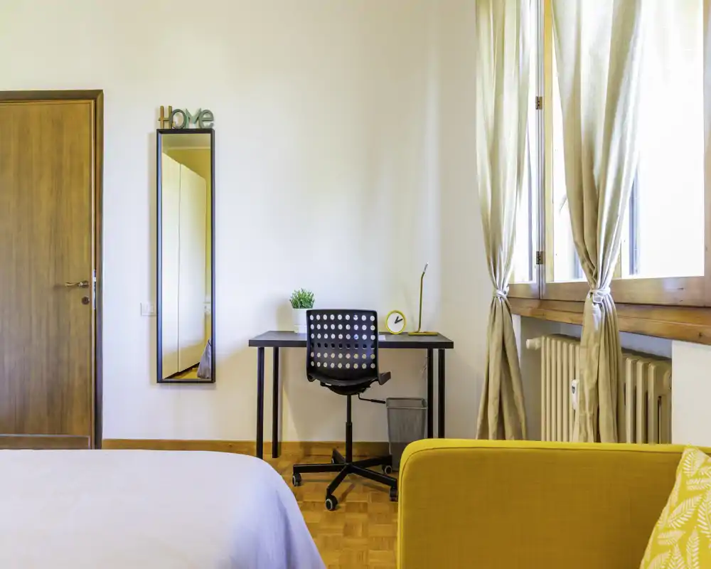 Camera da letto luminosa e accogliente con letto ben visibile, una scrivania e sedia vicino a una grande finestra, dettagli in legno caldo e un divanetto giallo che dona vivacità.