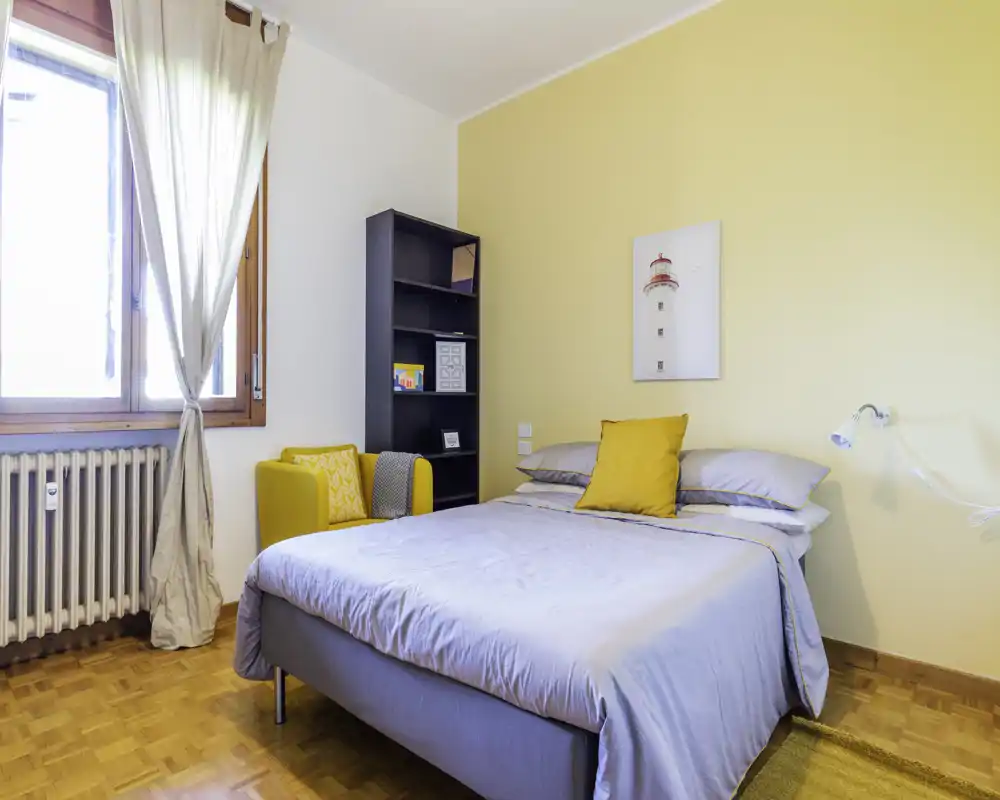 Camera da letto luminosa e curata, con letto matrimoniale in lenzuola morbide, poltrona gialla d'accento e tonalità calde alle pareti che la rendono accogliente.