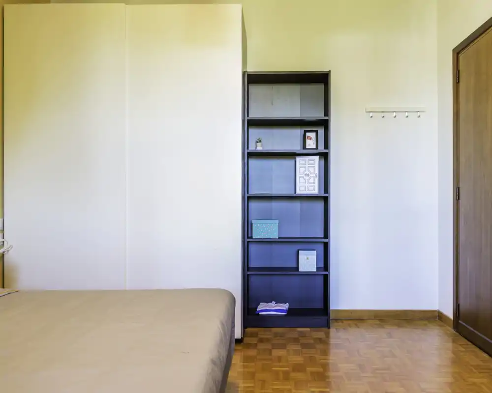 Angolo di camera da letto luminoso e arredato in modo semplice con letto visibile, armadio alto e mensole aperte — pavimento in parquet e pareti neutre per un ambiente tranquillo.