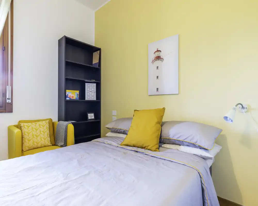 Camera da letto luminosa e ben arredata con letto matrimoniale rifinito, dettagli gialli caldi e una poltrona accogliente vicino alla finestra.