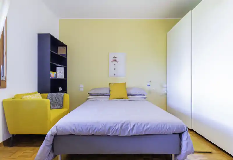 Camera da letto luminosa e ordinata con letto matrimoniale al centro, parete d'accento gialla e poltrona abbinata — ideale per un soggiorno accogliente.