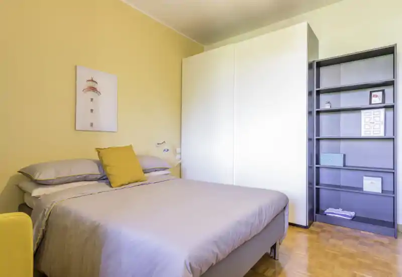 Camera da letto luminosa e ordinata con letto matrimoniale con biancheria neutra e cuscino giallo, grande armadio bianco e scaffale a vista; parete pastello calda che crea un’atmosfera accogliente.