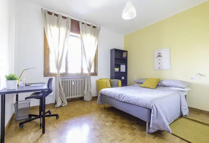 Camera da letto luminosa e moderna con letto matrimoniale, biancheria grigia, parete d'accento gialla e angolo studio vicino alla finestra. La luce naturale rende l'ambiente arioso e accogliente.