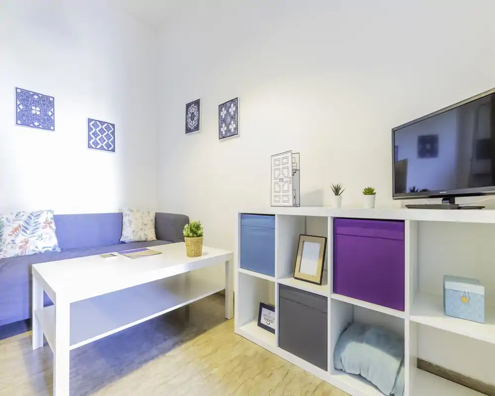 Zona giorno luminosa e moderna con divano compatto, tavolino e scaffalatura aperta con contenitori colorati; ambiente ordinato, ben illuminato e ben decorato.