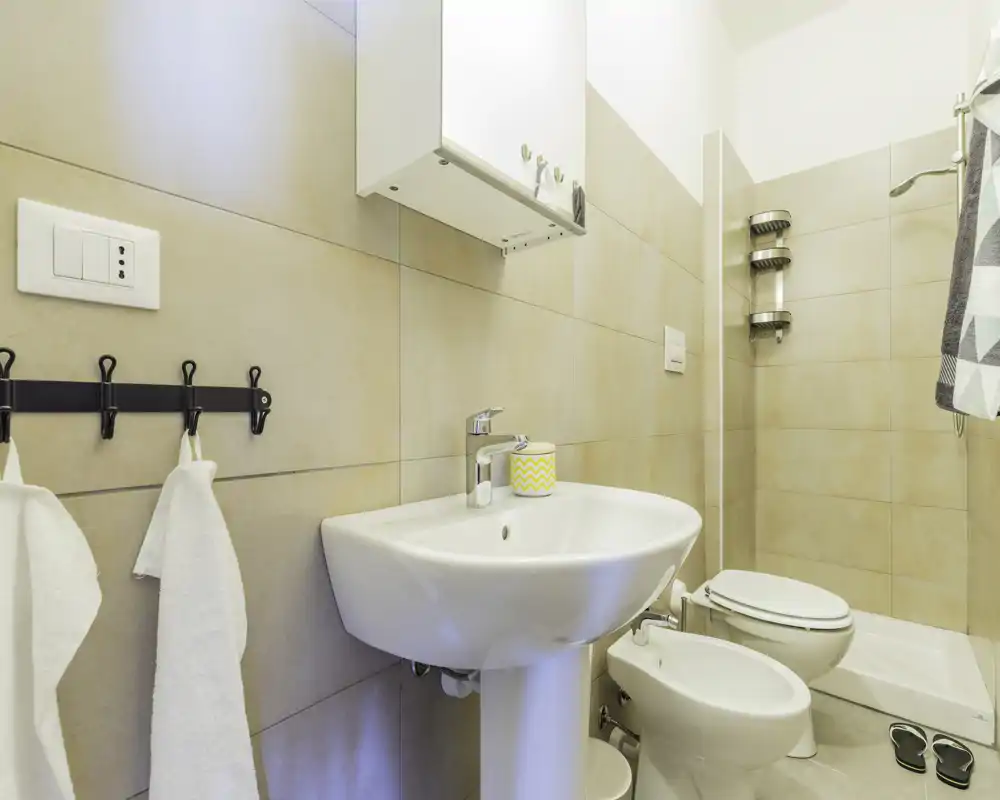 Bagno moderno e compatto con lavabo a colonna, bidet e WC, pareti piastrellate e doccia; ambiente luminoso e pulito con asciugamani e accessori minimal.