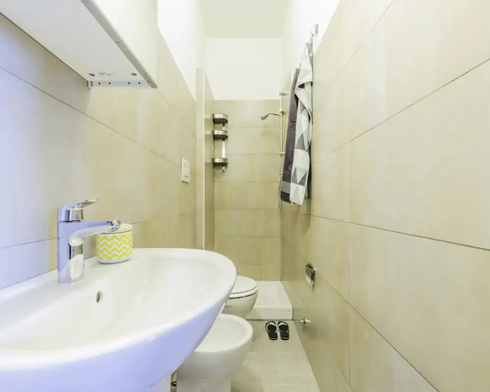 Bagno moderno e compatto, luminoso, con doccia a parete, lavabo, bidet e WC in piastrelle dai toni neutri.