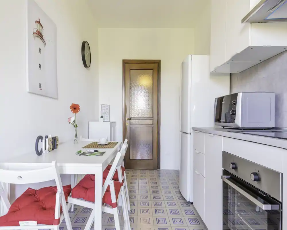 Cucina luminosa e compatta con pensili bianchi, forno incassato, microonde e un piccolo tavolo da pranzo con cuscini rossi — perfetta per un appartamento in città.