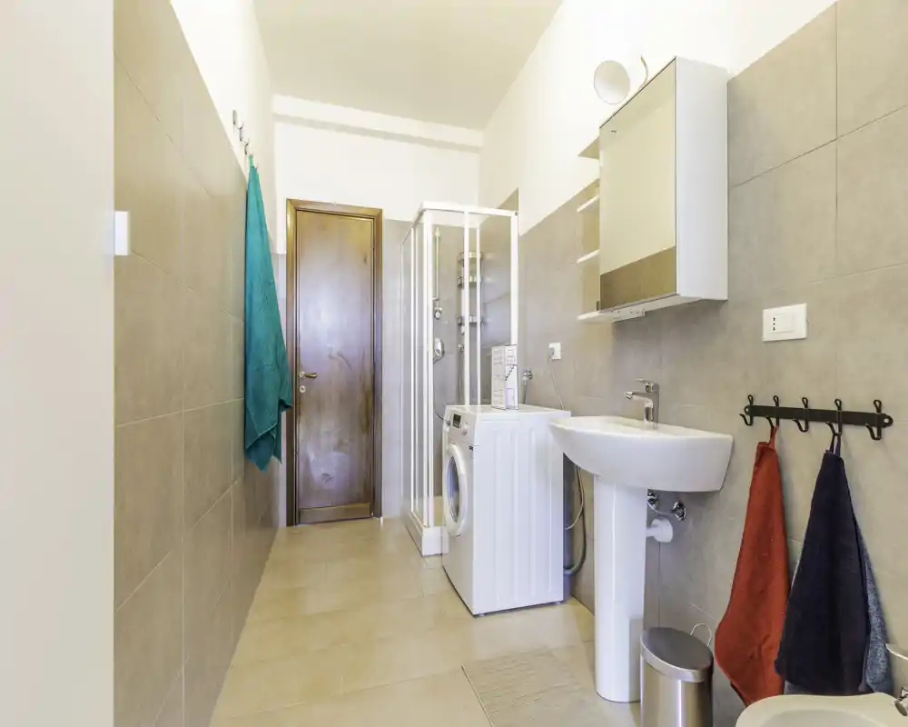 Bagno moderno e luminoso con cabina doccia, lavabo a piantana, pensile specchio e lavatrice. Piastrelle neutre e arredi ordinati creano uno spazio pratico e pulito.