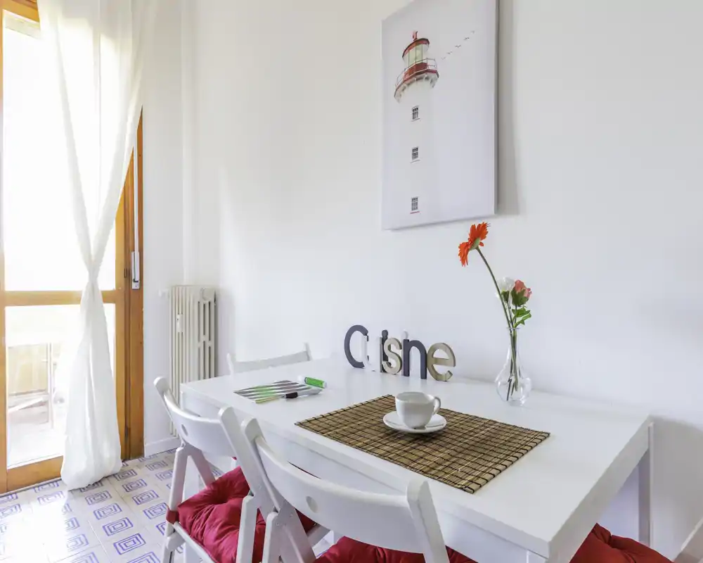 Accogliente angolo pranzo luminoso con tavolo bianco, sedie richiudibili con cuscini rossi e dettagli decorativi come vaso con fiori e quadro — perfetto per pasti informali.
