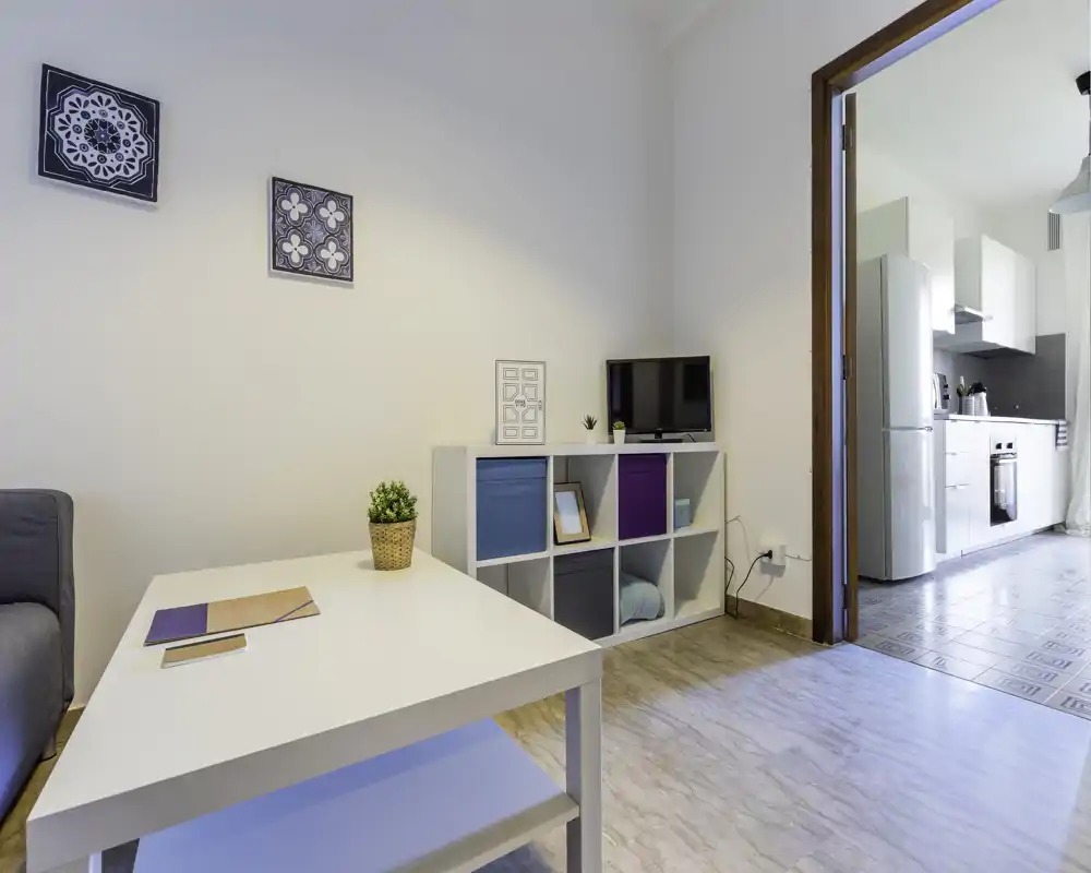Zona giorno luminosa e moderna con tavolino bianco, mobiletto a cubi, piccola TV e pavimento chiaro; vista aperta su una cucina contemporanea e pulita.