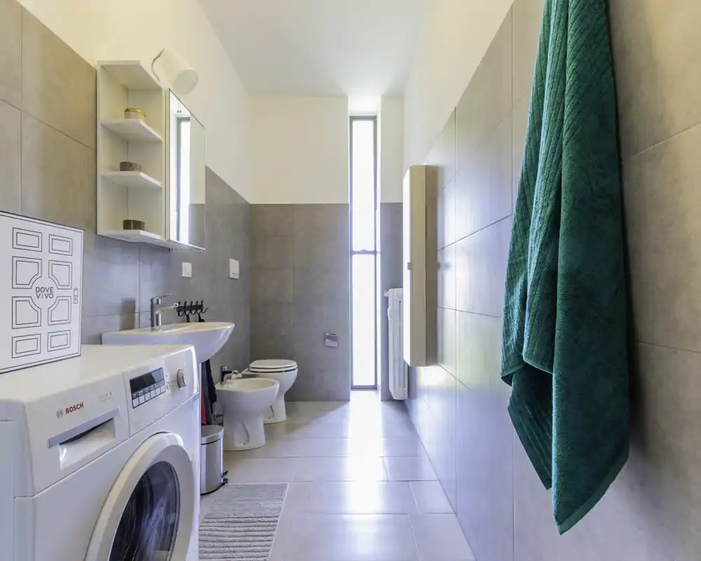 Bagno moderno e luminoso con lavatrice, lavabo, bidet e WC; una finestra verticale assicura luce naturale e piastrelle dalle pareti donano un aspetto pulito.