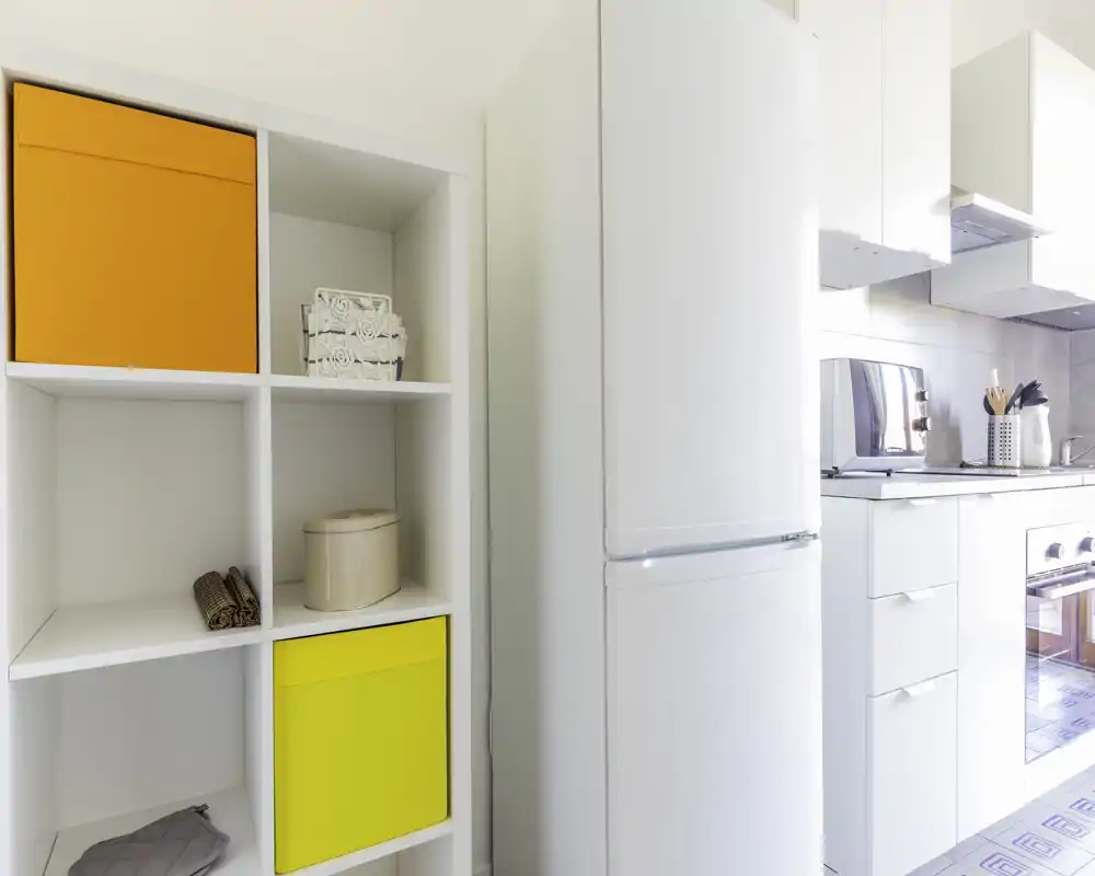 Angolo cucina moderno e luminoso con frigorifero alto bianco, forno incassato e mobili bianchi puliti; contenitori colorati aggiungono un tocco vivace.