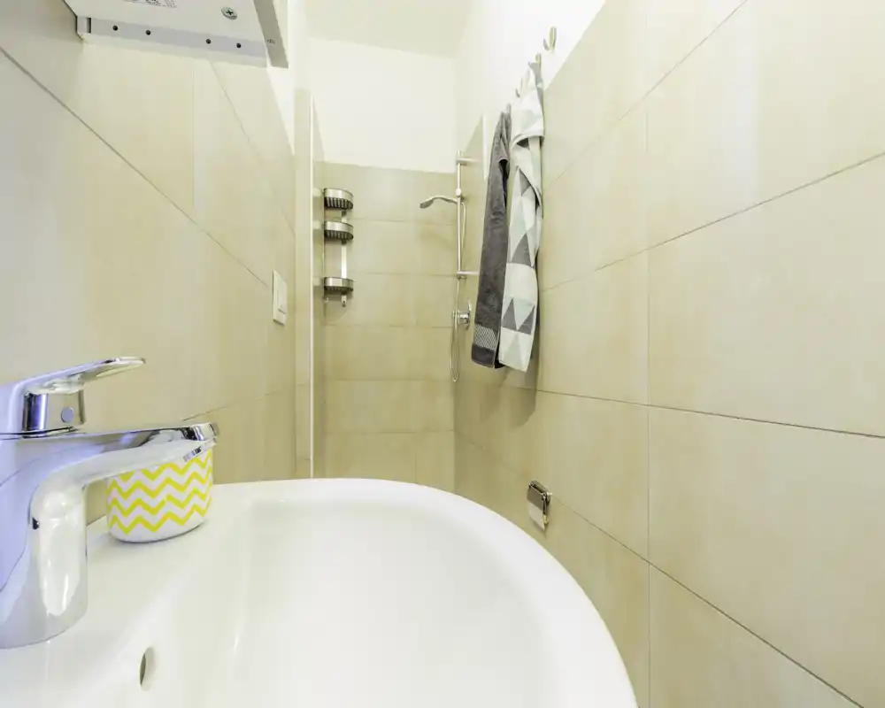 Bagno moderno compatto con lavabo bianco, rubinetto cromato e doccia walk-in in piastrelle neutre; asciugamani appesi donano un aspetto pulito e luminoso.