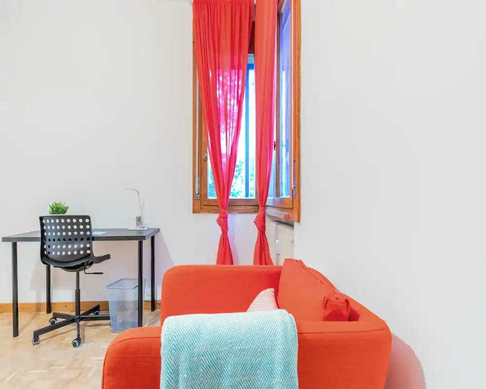 Bright, modern living area with a vibrant orange sofa, a small desk and a window dressed with red curtains — a compact, well-lit space ideal for relaxing or light work.