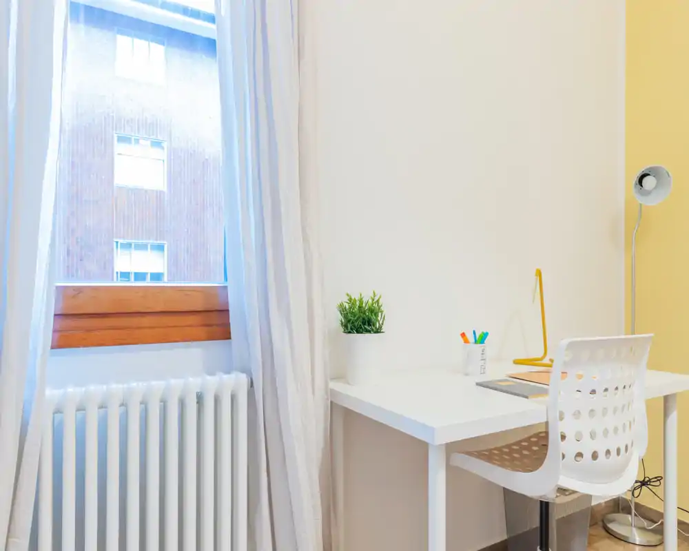 Bright, minimalist home-office corner beside a large window with a white desk, modern chair, desk lamp and a small plant — ideal for focused remote work.