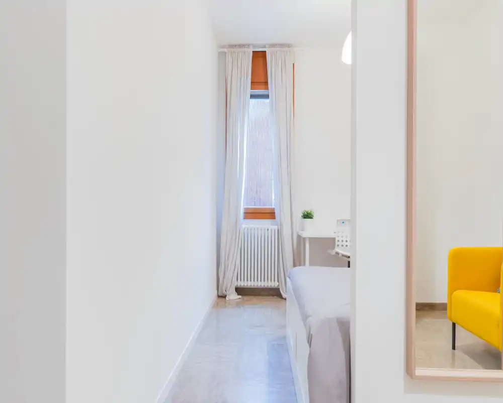 Bright minimal bedroom corner with a single bed, white walls, a window with light curtains and a small desk—clean, airy and simply furnished.
