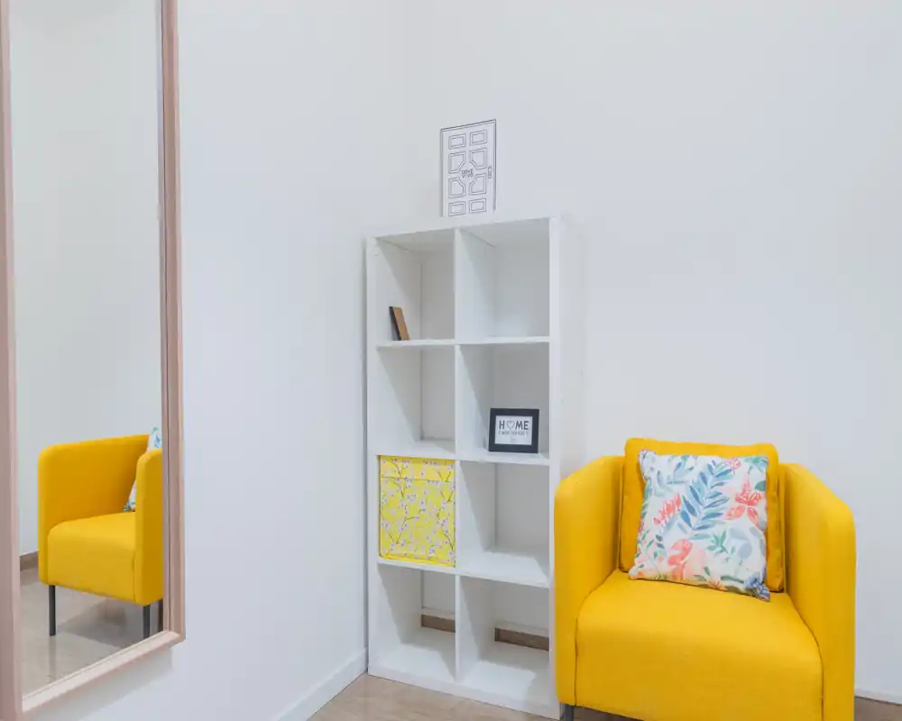 Bright, minimalist living area with a cheerful yellow armchair, decorative cushion and white shelving; clean walls and a large mirror create a sense of space.
