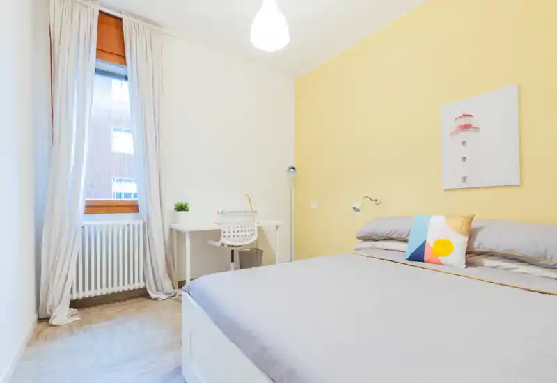Bright, modern bedroom with a comfortable double bed, soft gray bedding, a decorative cushion, and a small desk by the window — ideal for rest and light work.