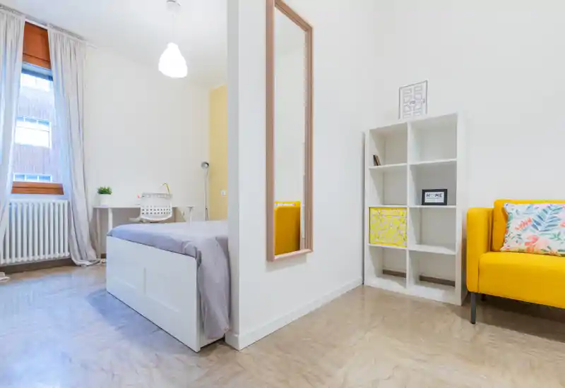 Bright, modern bedroom with a comfortable single/double bed, a desk by the window and a cheerful yellow accent sofa. Clean minimalist styling and natural light make this room ideal for studying and relaxing.