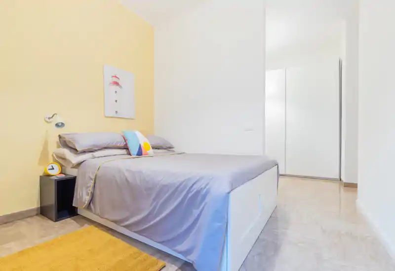 Bright, modern bedroom with a full bed, soft grey linens, decorative cushion and a small bedside table; clean lines and warm yellow accent wall create a welcoming atmosphere.