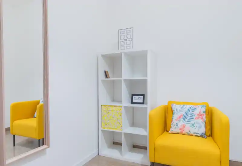 Bright, minimalist living area with a cheerful yellow armchair, decorative cushion and white shelving; clean walls and a large mirror create a sense of space.