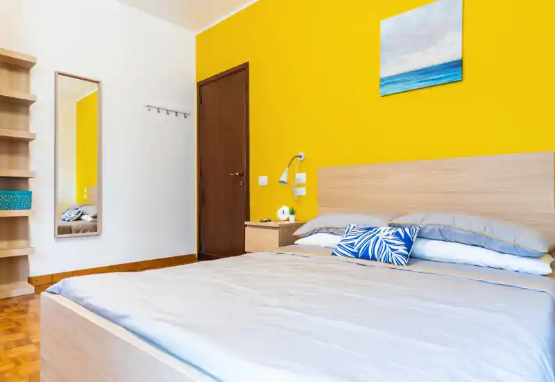 Bright, modern bedroom with a large double bed, wooden headboard and bedside table. The yellow accent wall and decorative cushions create a cheerful, welcoming atmosphere ideal for rental listings.