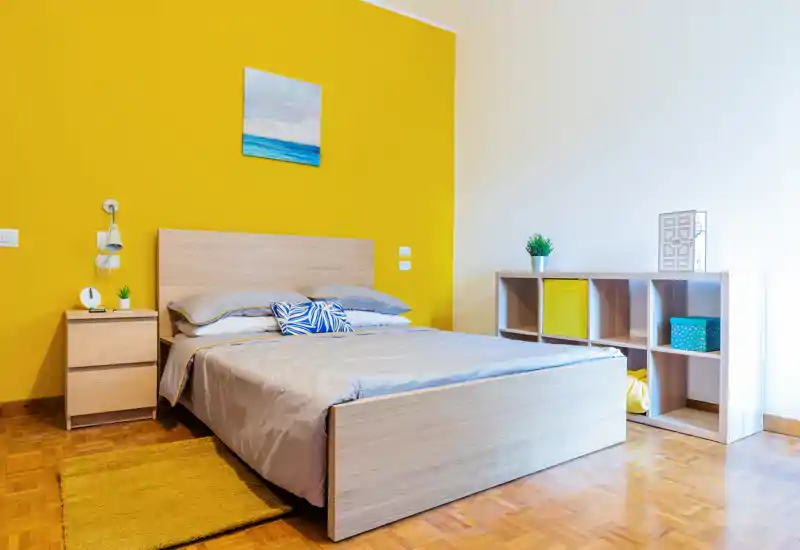 Bright, modern bedroom with a wooden double bed against a bold yellow accent wall, bedside table, decorative cushions and a low cube shelving unit. The room feels fresh and well-lit, with simple Scandinavian-style furnishings.