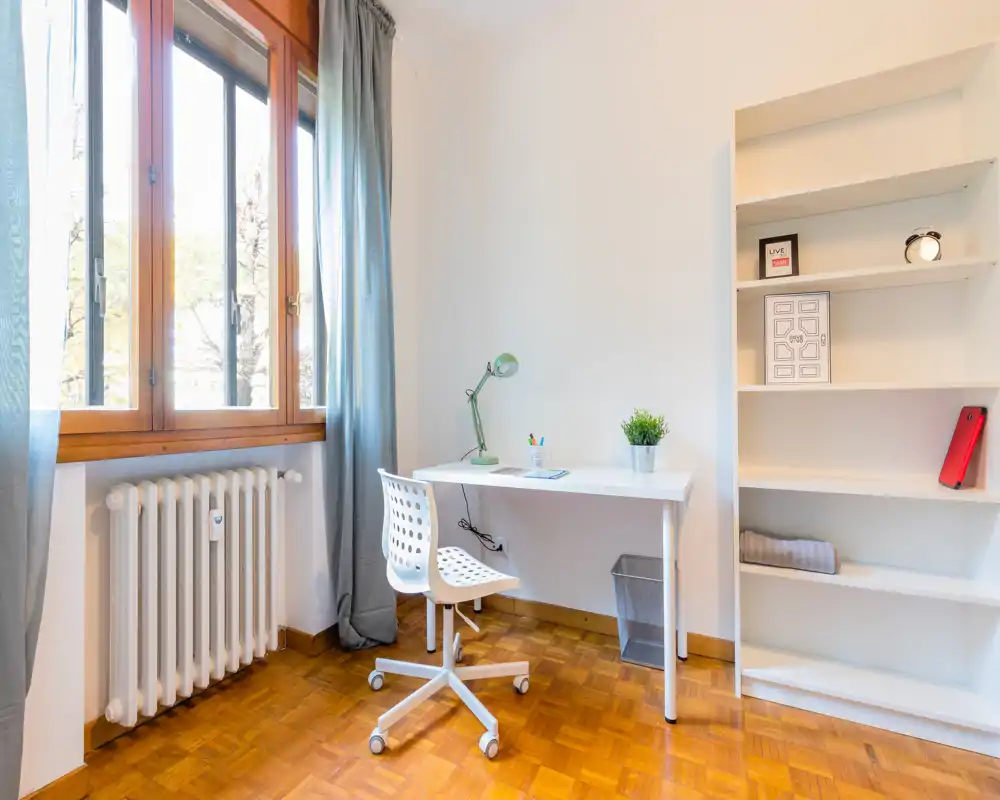 Bright, minimal home workspace with a white desk, swivel chair, lamp and shelving next to a large window with curtains and a radiator.