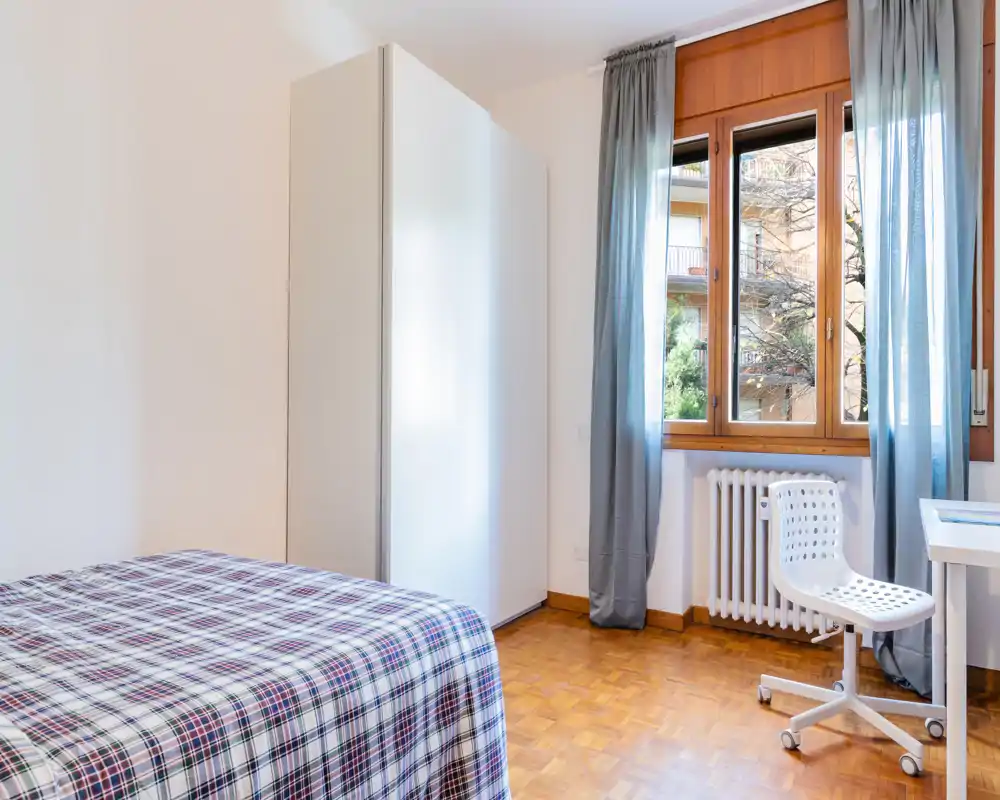 Bright, simply furnished bedroom with a patterned bedspread, white wardrobe and a window letting in natural light — ideal for rental listings seeking a clean, airy sleeping space.
