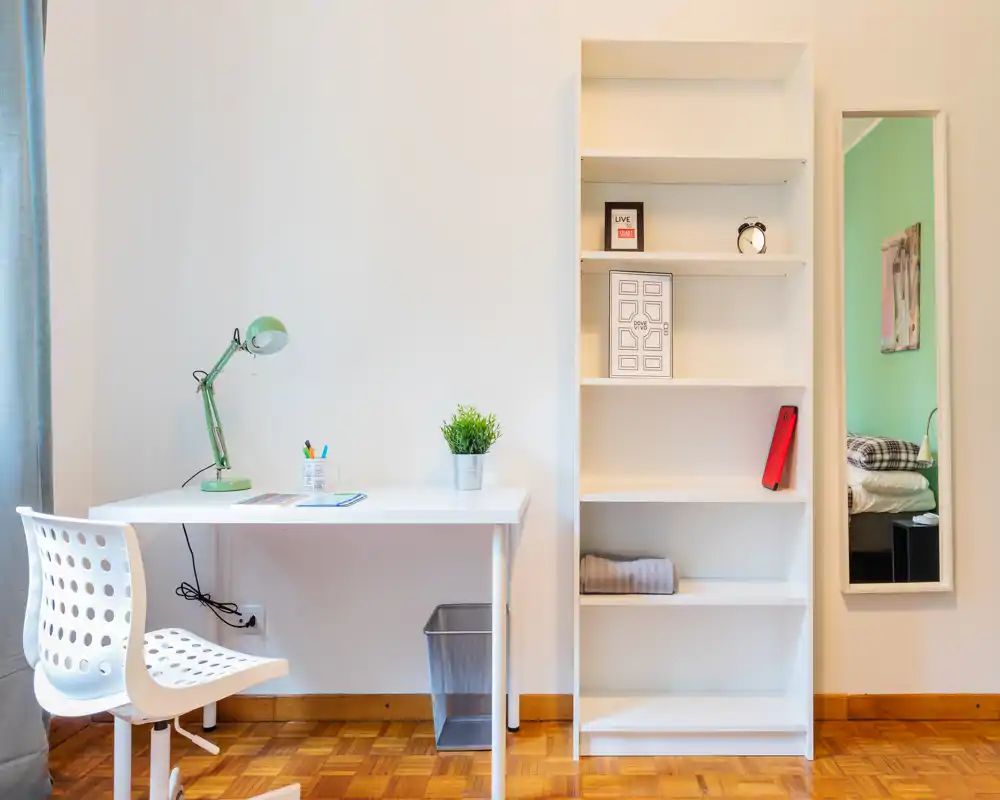 Bright, minimal bedroom corner with a study desk, shelving and a mirror that shows the bed in reflection. Clean white furniture and wooden flooring create a simple, functional space ideal for solo stays.