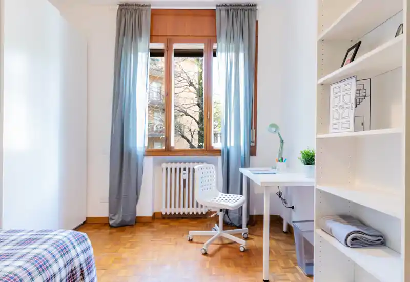Bright, minimal bedroom with a single bed, desk and shelving. Large window provides natural light and a clean parquet floor gives a warm touch.