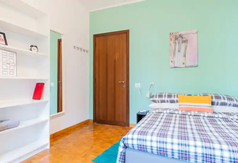 Bright, neatly styled bedroom with a double bed dressed in plaid linens, accent pillows and a bedside lamp; mint-green accent wall and wooden door add a fresh, cozy atmosphere.