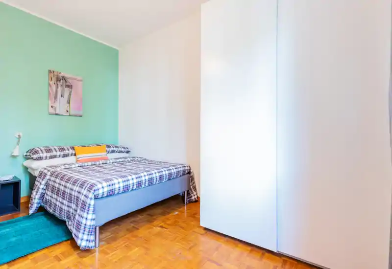 Bright, tidy bedroom with a double bed dressed in plaid bedding, a colorful accent pillow and a large white wardrobe. Soft mint green accent wall and wooden parquet flooring create a fresh, welcoming atmosphere.